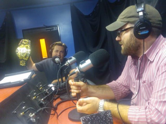 Two hosts at the microphones, one holding up a championship belt