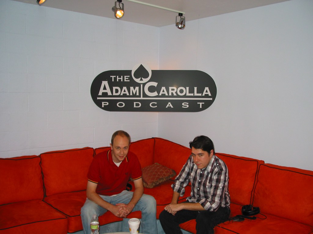 Marc and Tod on a red sectional couch beneath a large 'The Adam Carolla Podcast' logo on a white brick wall